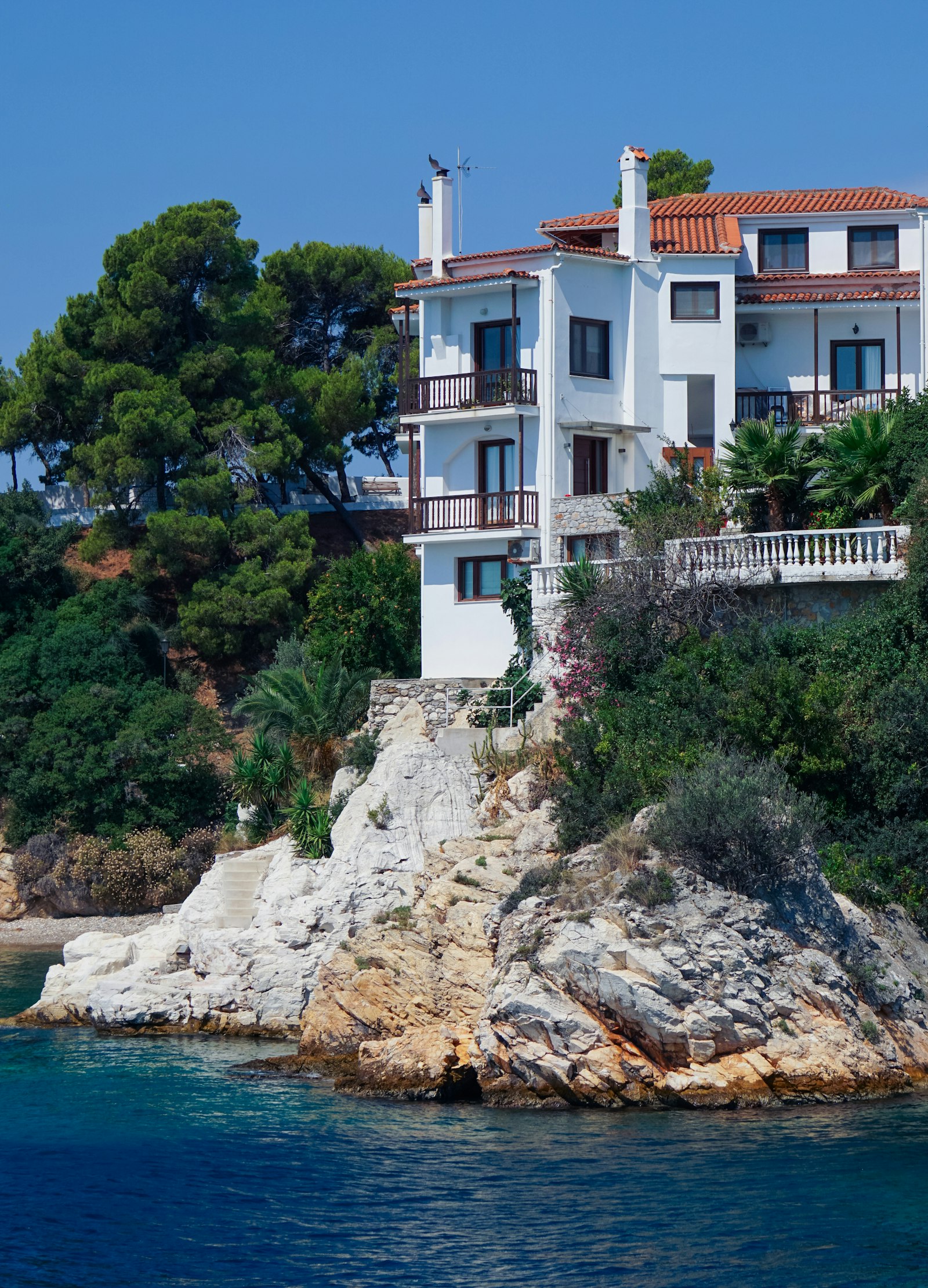 A white coastal house with timber shutters on a rocky cliff above the Mediterranean.